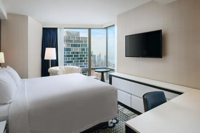 A hotel bedroom with a king bed, a white armchair, a wall-mounted television and a desk, with floor-to-ceiling windows showing city buildings.