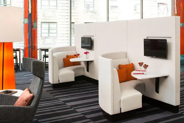 A lounge with white upholstered booths, wall-mounted televisions, orange cushions and a grey armchair beside large windows.