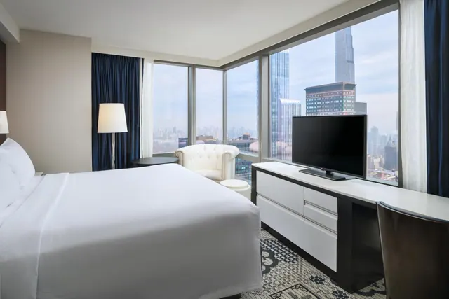 A bedroom with a king bed, corner floor-to-ceiling windows overlooking city skyscrapers, a flat-screen TV on a white dresser, and a white armchair by the window.