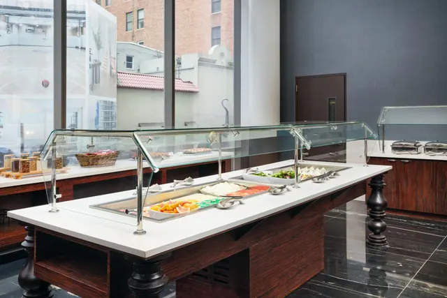 A buffet counter with glass sneeze guards holds fruit, salad and other dishes, with tall windows overlooking nearby brick buildings.
