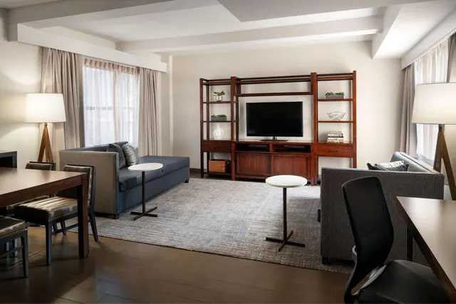 A living room with a dark blue sofa, a grey sofa, round side tables, a wooden shelving unit holding a flat-screen TV, and a dining table and desk chair in the foreground.
