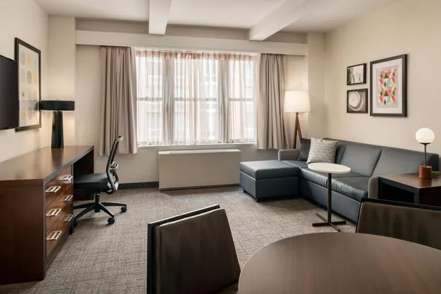 A hotel suite living area with a grey sectional sofa, a round side table, a wooden desk with an office chair, and a window with sheer curtains.