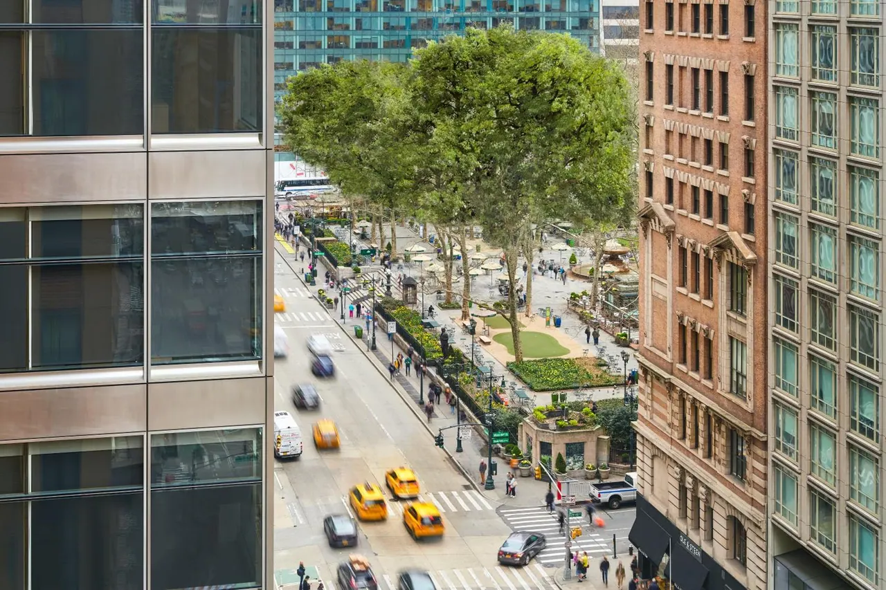An elevated view down onto a city street with yellow taxis, a tree-lined plaza with a lawn and umbrellas, and brick and glass buildings on either side.