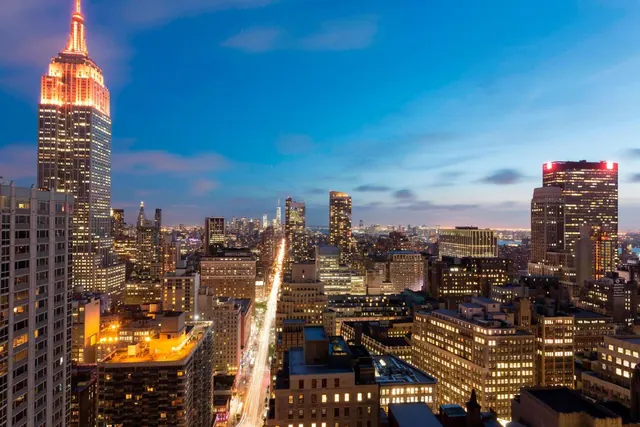 Twilight aerial view over Manhattan toward the illuminated Empire State Building, with lit office towers and light trails on a street below.