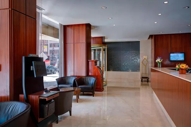 A hotel lobby with leather armchairs, a small coffee table, a computer kiosk, a reception desk with flower arrangements, and a street with a parked car visible through the front windows.
