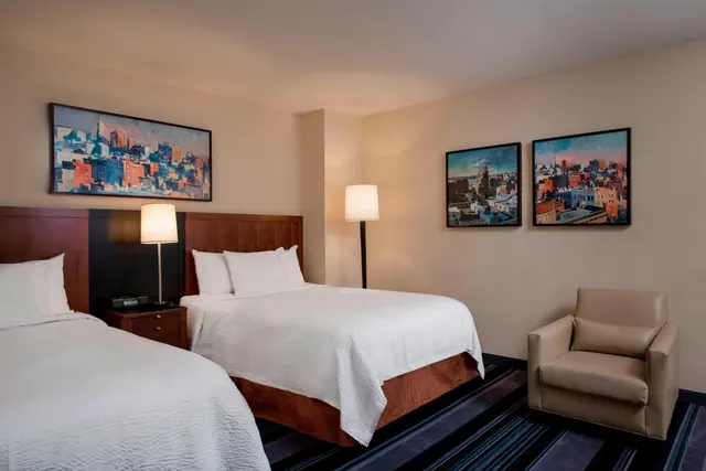 A hotel bedroom with double beds under white bedding, bedside lamps, framed city paintings on the walls, and an upholstered armchair in the corner.