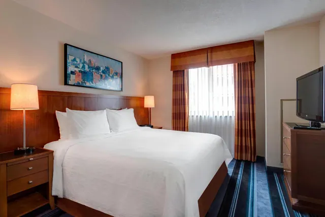 A made king bed with white linens sits beside a wooden nightstand and lamp, with a wall-mounted television on a dresser opposite and a window with striped drapes behind.