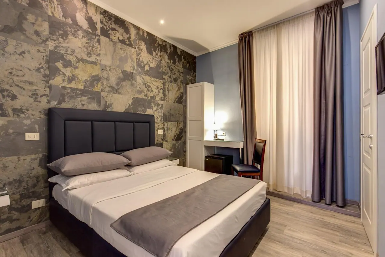 A double bed with a dark upholstered headboard stands against a stone-patterned wall, beside a white wardrobe, a desk with a chair, and a minibar.