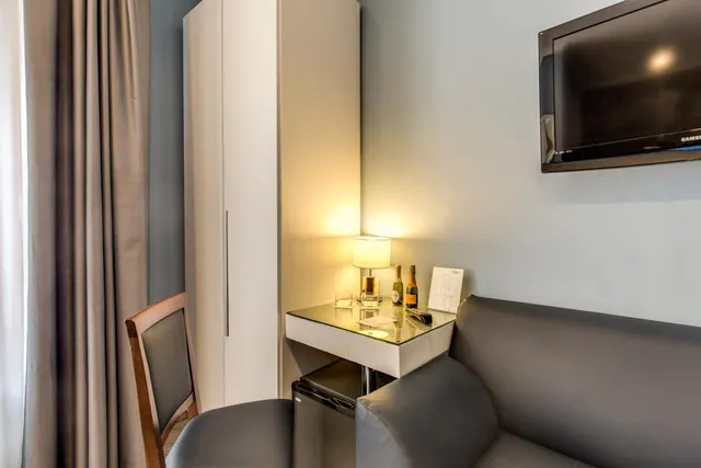 A corner of a hotel room with a wall-mounted flat-screen television, a grey sofa, a glass-topped table with a lamp and two bottles, and a tall white wardrobe.