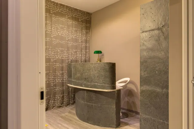 A curved stone-clad reception desk with a green banker's lamp and a white swivel stool stands in front of a wall covered in script-patterned fabric.