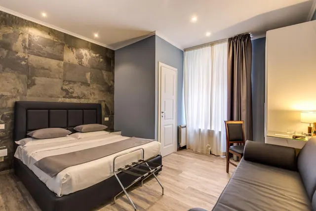 A bedroom with a double bed, a dark stone-effect accent wall, a leather sofa, a white wardrobe, a desk lamp and a window with sheer curtains.