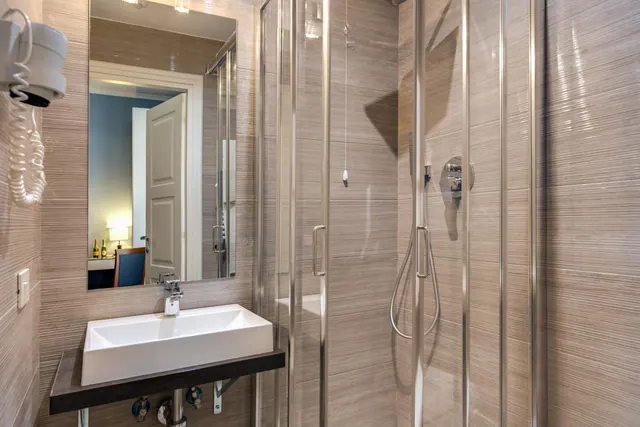 A bathroom with a wall-mounted basin, a glass-enclosed shower, and a mirror reflecting a bedroom doorway.