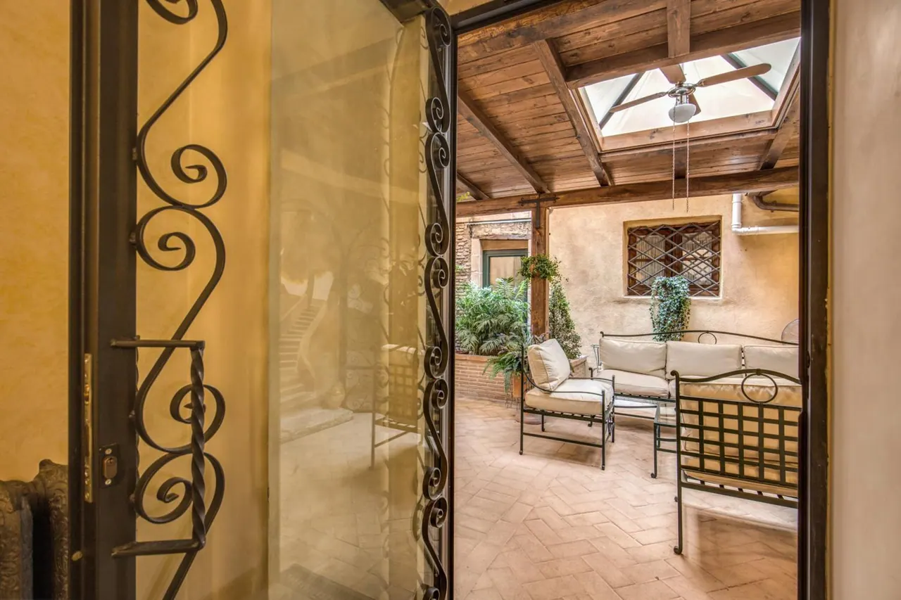An open wrought-iron gate leads into a covered terrace with a wooden beamed ceiling, a skylight with a ceiling fan, white-cushioned metal sofas and chairs, and a terracotta herringbone floor.