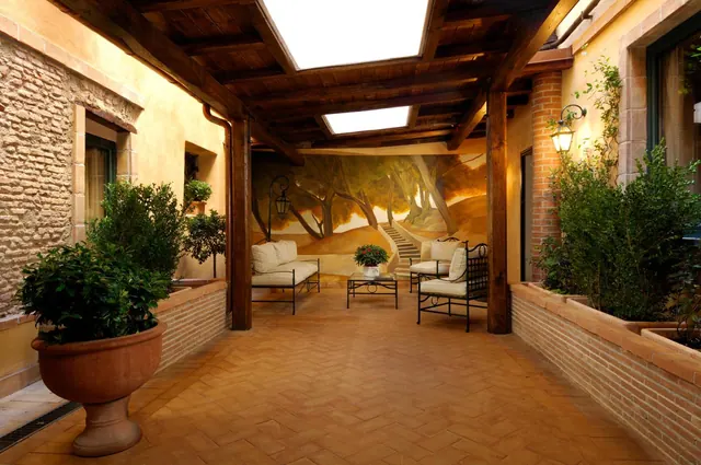 A covered terracotta-tiled patio with wooden roof beams, a landscape mural on the back wall, potted plants and a seating area with a sofa and armchairs.