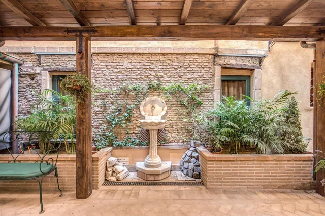 A covered courtyard with a stone wall fountain, potted palms, climbing ivy on brick, and a green metal bench under wooden beams.