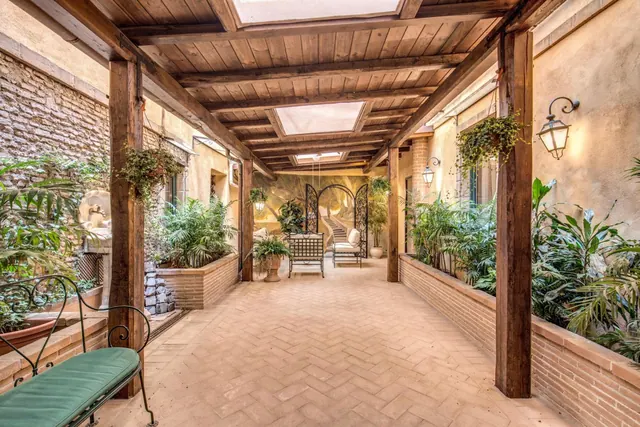 A covered brick-paved passage with wooden beams, skylights, potted plants along both sides and a wrought iron bench with a green cushion in the foreground.