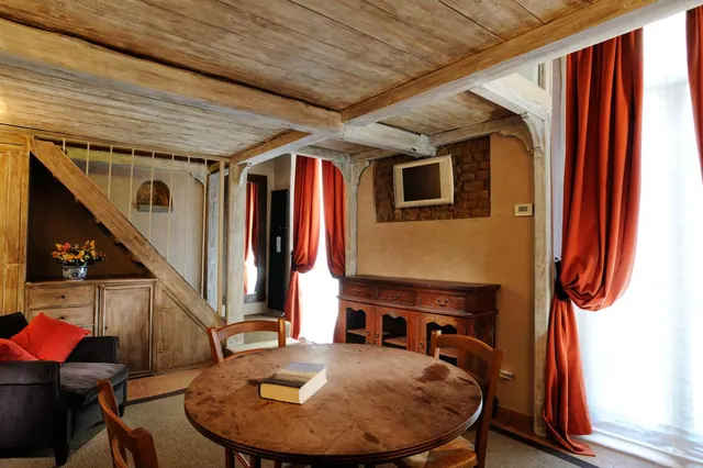 A living area with a grey sofa and red cushions, a round wooden dining table with a book, a wooden sideboard, a wall-mounted television, and red curtains beside tall windows under an exposed timber ceiling.