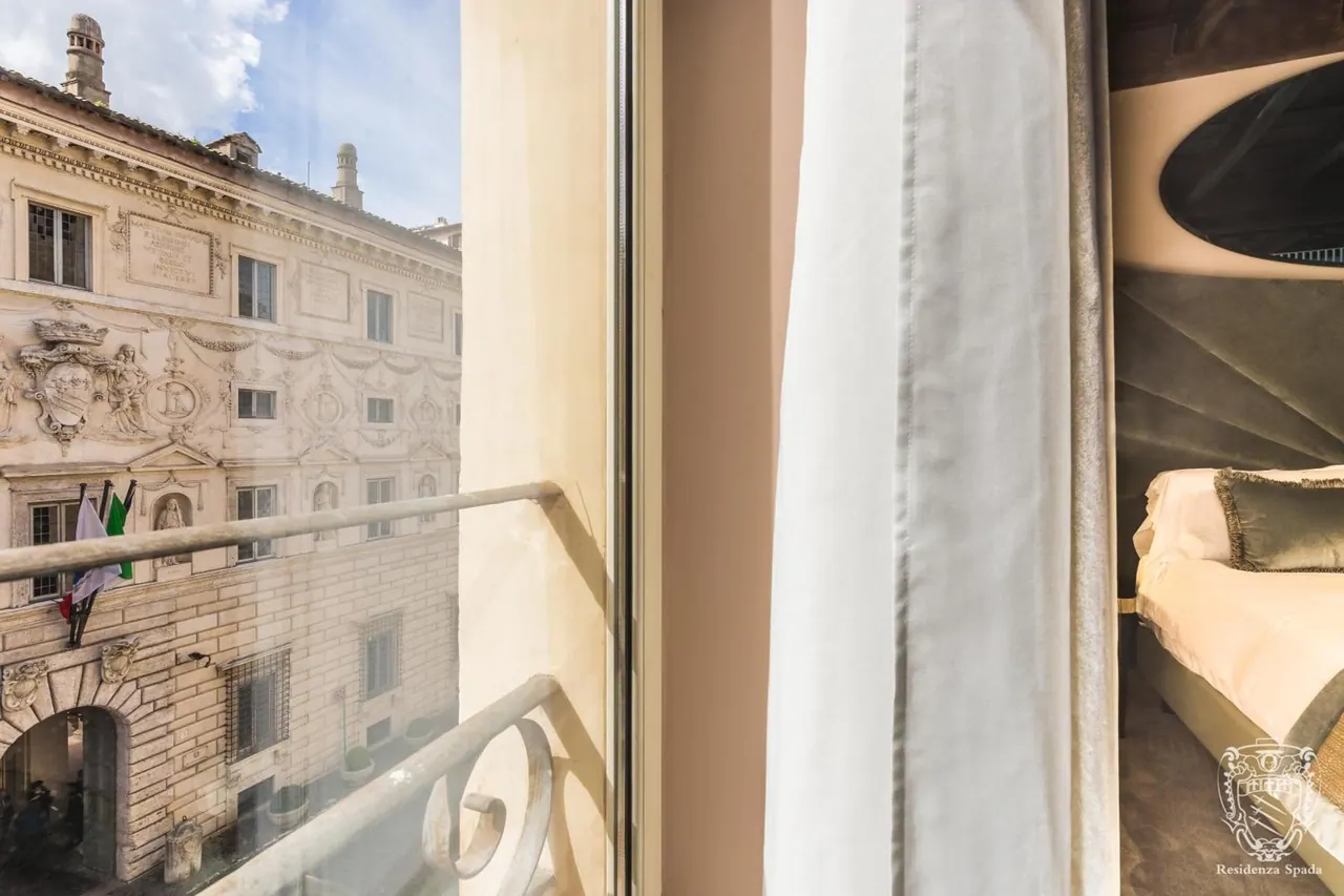 Looking out from a room window and balcony railing onto an ornate stone palazzo facade with flags, with a made double bed visible inside on the right.