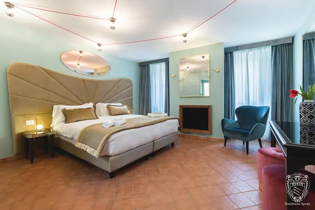 A bedroom with a large upholstered headboard and double bed, a teal armchair by the window, and terracotta floor tiles, with a Residenza Spada logo watermark in the corner.