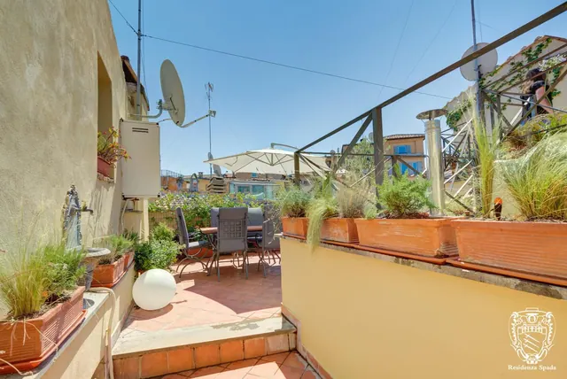 A rooftop terrace with a table, chairs, a white parasol, potted plants and a view over neighbouring rooftops and buildings, with a Residenza Spada watermark in the corner.