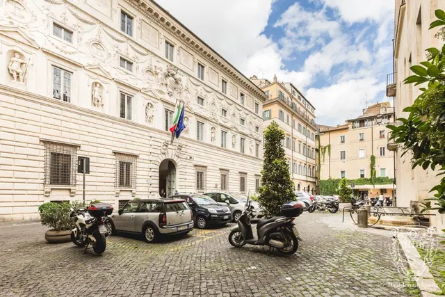 The ornate stone facade of a historic palazzo with an arched entrance and flags overlooks a cobbled square where cars and scooters are parked.