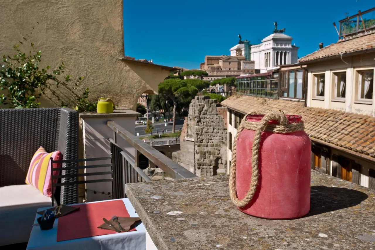 A rooftop terrace with wicker seating and a set table, with a red lantern on a stone ledge, looking out over Rome rooftops, ancient ruins and the Vittoriano monument.