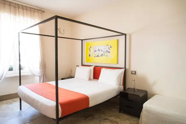 A bedroom with a black four-poster metal bed with white linens and an orange throw, a yellow painting on the wall, a dark nightstand, and a window with sheer curtains on the left.