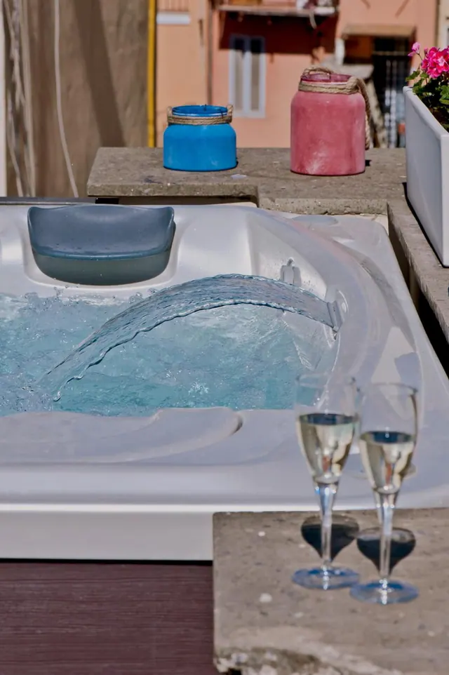 An outdoor hot tub with a jet of water flowing in, with two champagne glasses on a concrete ledge and coloured pots and a flowering planter behind.