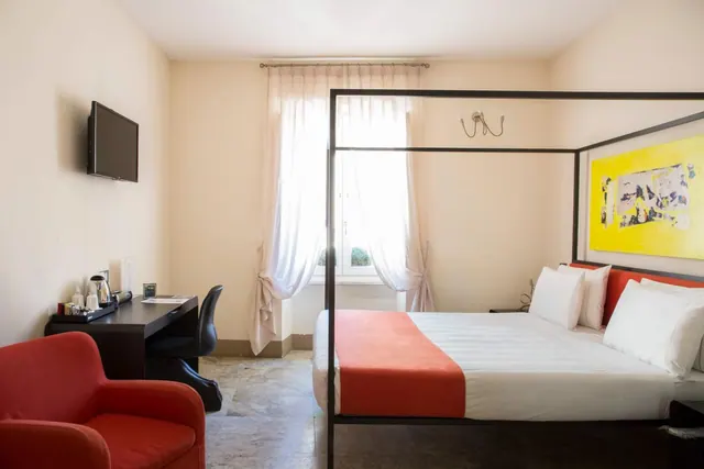 A bedroom with a four-poster double bed, a red armchair, a wall-mounted television, a desk with a kettle, and a window with sheer curtains.