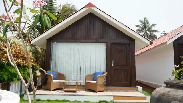 A tropical cottage exterior has a covered sitting area with two chairs and a small table surrounded by greenery.