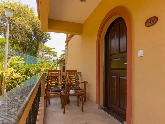 A furnished private balcony overlooks lush tropical greenery beside an arched wooden hotel room door.