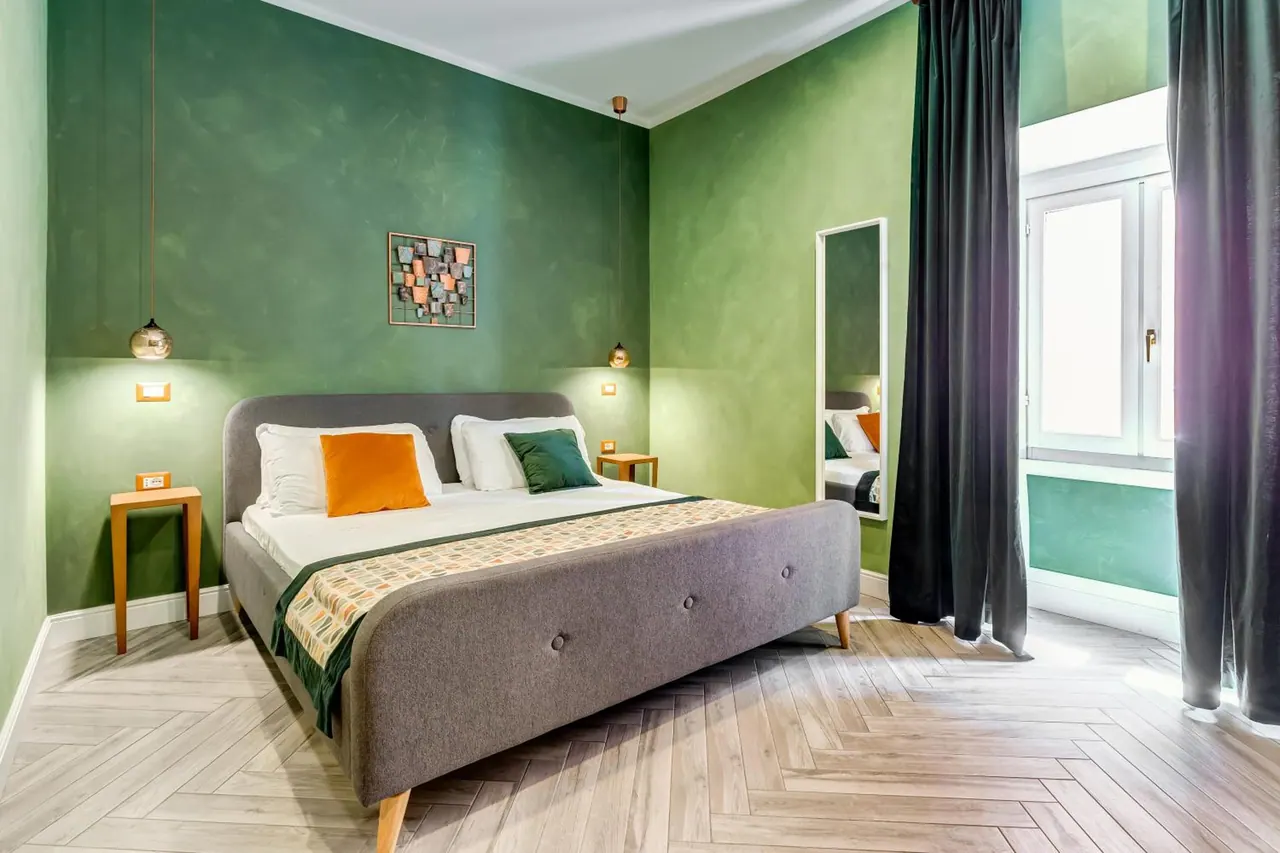 A bedroom with green textured walls, a grey upholstered double bed with orange and green pillows, pendant lamps, a tall mirror and dark curtains beside a window.
