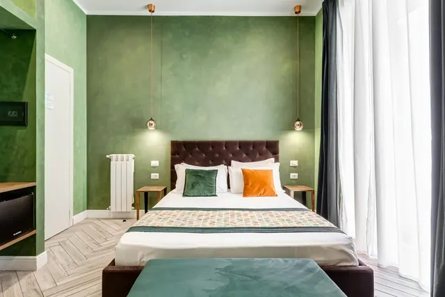 A double bed with green and orange cushions sits against a green textured wall beside a window with sheer white curtains.