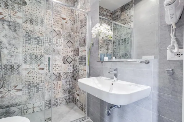 A bathroom with a glass-enclosed walk-in shower, patterned tiled walls, a wall-mounted basin, a mirror and a white orchid on the wall.