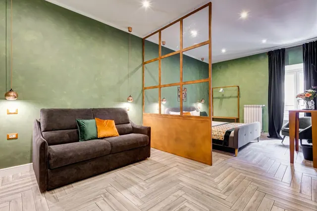 A studio room with a dark brown sofa with green and orange cushions in front of a green wall, with a glass and brass partition and a bed visible behind it.