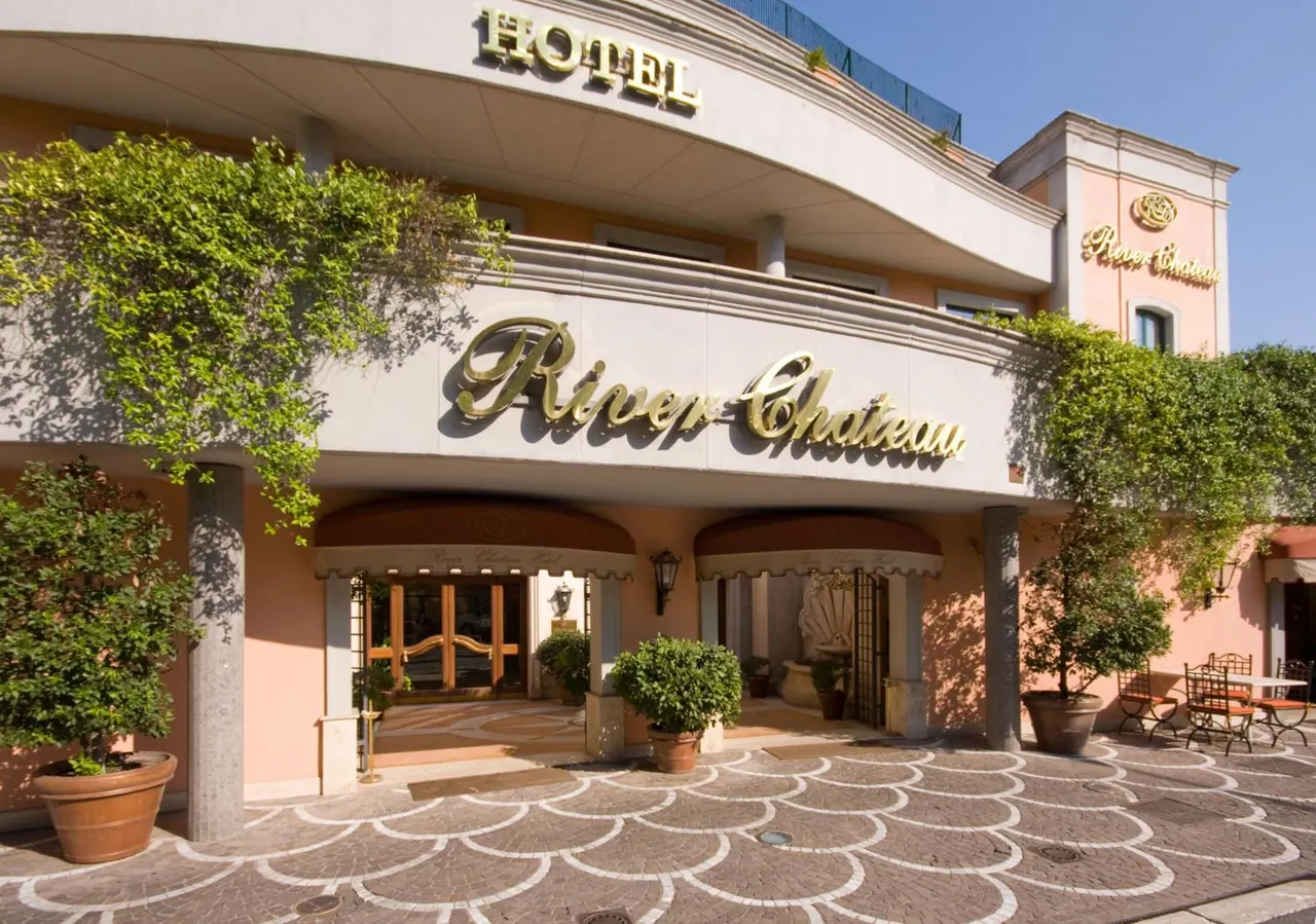 The entrance facade of the River Chateau hotel, with gold signage, potted plants and a patterned stone forecourt under clear daylight.