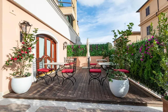 A wooden deck terrace with wrought iron tables, red-cushioned chairs, white planters and flowering plants beside a pale pink building.
