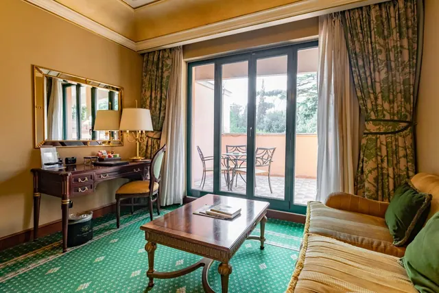 A hotel sitting room with a wooden desk, a sofa and coffee table, and glass doors opening onto a terrace with metal chairs and a table.