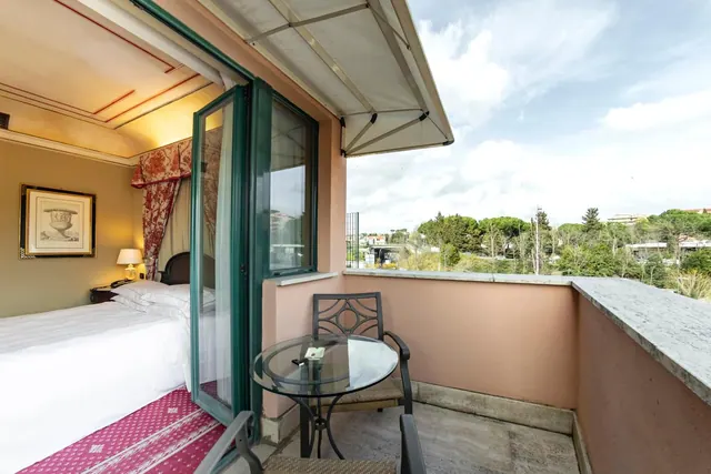 A balcony with a glass table and metal chair opens through glass doors to a bedroom with a made white bed, and trees and buildings are visible beyond the railing.