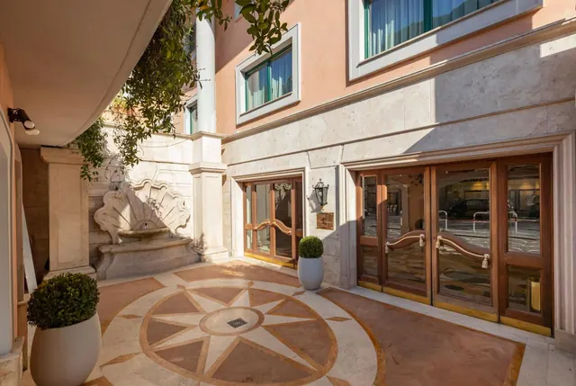 Entrance courtyard with a marble compass-rose floor, a wall fountain, potted topiary plants and wooden glass entrance doors.