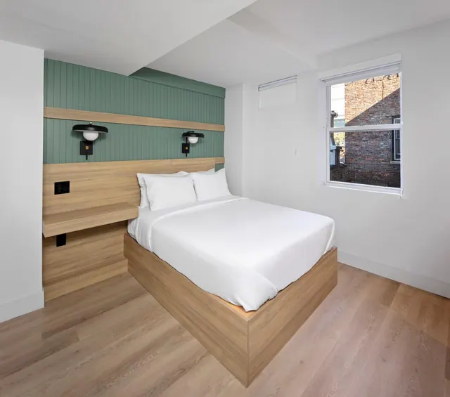 A bedroom with a double bed, wall lamps on a green panelled wall, and a window looking onto a brick building outside.