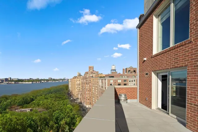 A tiled terrace with a grey parapet overlooks a river, a green park and brick apartment buildings under a blue sky.