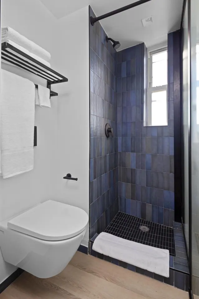 A bathroom with a walk-in shower tiled in dark blue, a wall-mounted toilet, and folded white towels on a black rack.