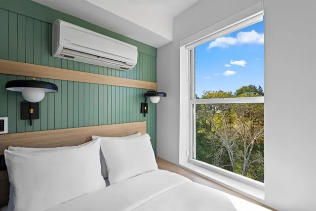 A double bed with white linens sits beside a window that looks out onto trees and a blue sky, with a wall-mounted air conditioner above the bed.