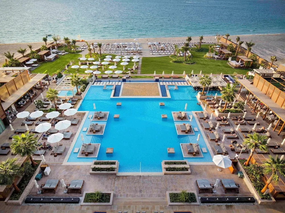 Aerial view of a large swimming pool with white umbrellas and sun loungers, with a sandy beach and the sea beyond.