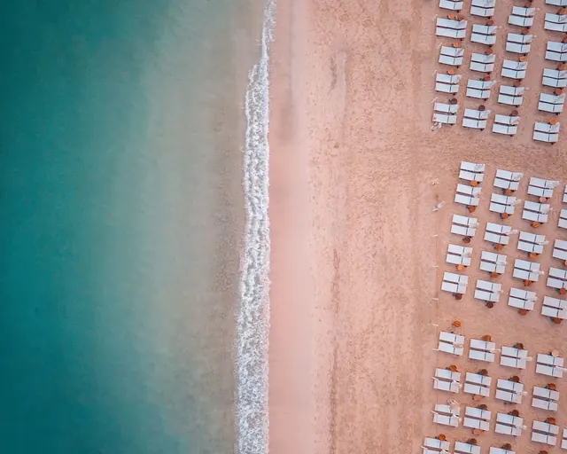 Aerial view of rows of white sun loungers and parasols on a sandy beach beside turquoise sea.