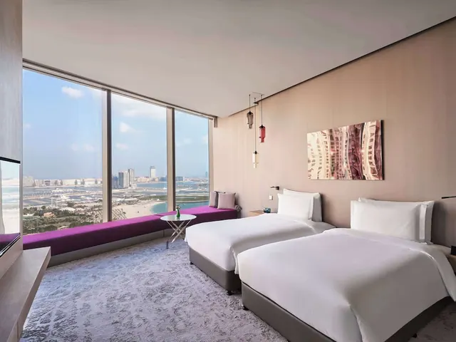 A bedroom with two white beds, a purple window bench, and floor-to-ceiling windows overlooking a city skyline and the sea.