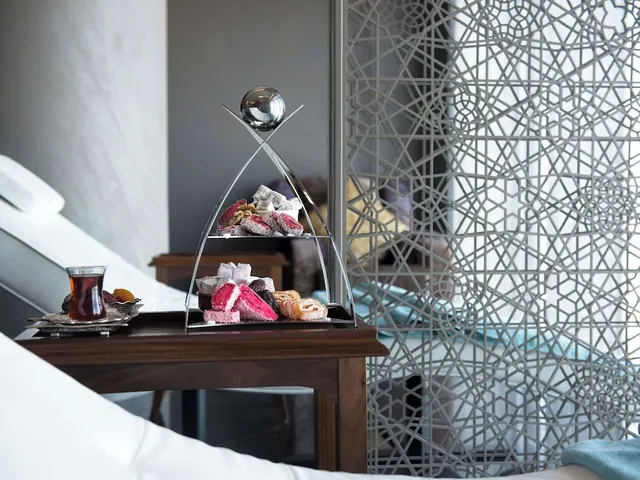 A bedroom with white bedding, a wooden side table holding a tiered tray of sweets, a glass of tea and dates, and a decorative metal lattice screen beside it.