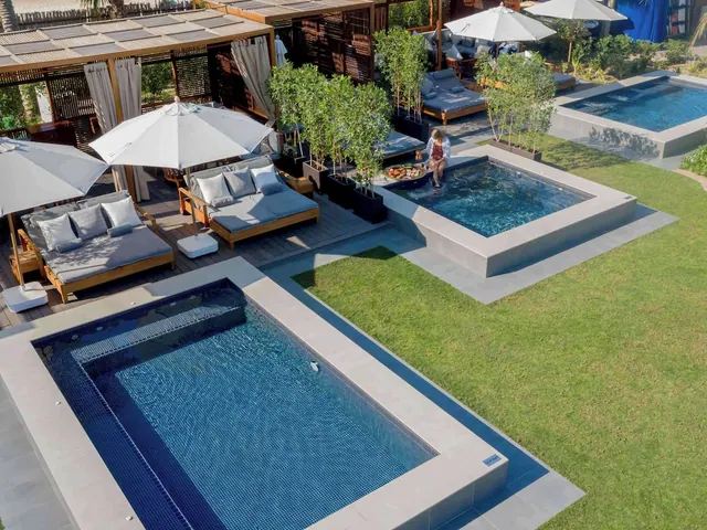 Elevated view of a large rectangular pool and a small square pool beside daybeds, white parasol umbrellas and a lawn, with one woman seated at the edge of the small pool.