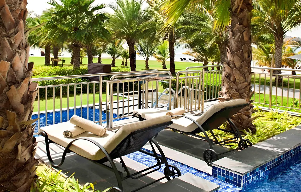 Two cushioned sun loungers with folded towels stand on a poolside deck beside a blue-tiled pool edge, with palm trees and a lawn visible beyond the metal railing.